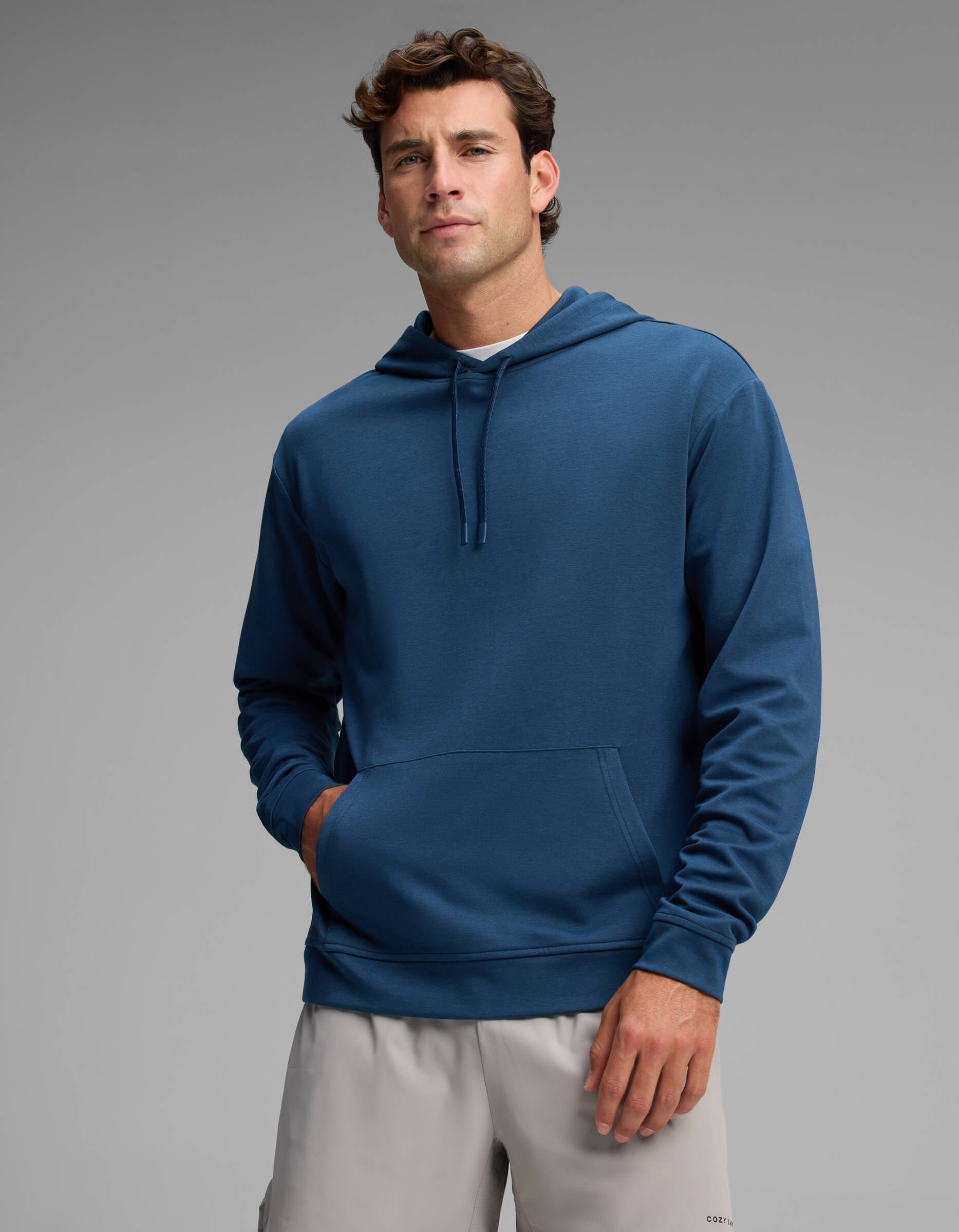 A man wearing the Cozy Earth Men's Versa Hoodie in blue and light gray shorts stands with one hand in his pocket against a plain gray background.|Color:Obsidian
