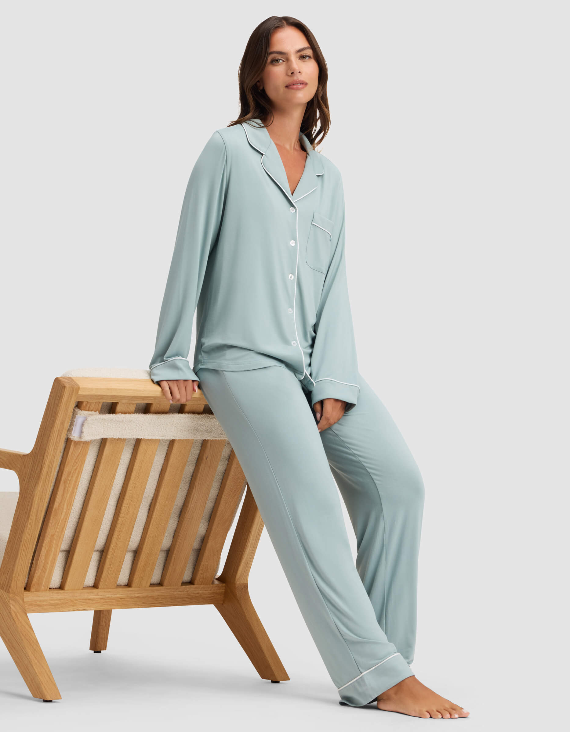 A woman in the Cozy Earth Women's Bamboo Stretch-Knit Long Sleeve Pajama Set in light blue sits casually on the arm of a wooden lounge chair with a neutral cushion, set against a plain light background. |Color:Brook