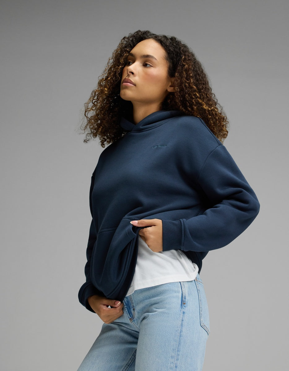Wearing the Cozy Earth Women’s CityScape Hoodie in Eclipse over a white shirt and light blue jeans, a person with curly hair stands against a plain gray background, gazing slightly upward.|Color:Eclipse