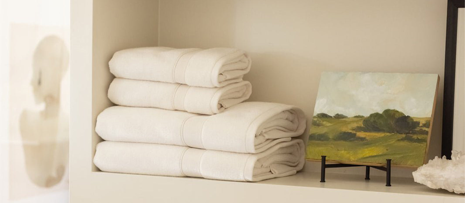 Stack of Cozy Earth Luxe Bath Towels sit on a shelf next to a landscape painting