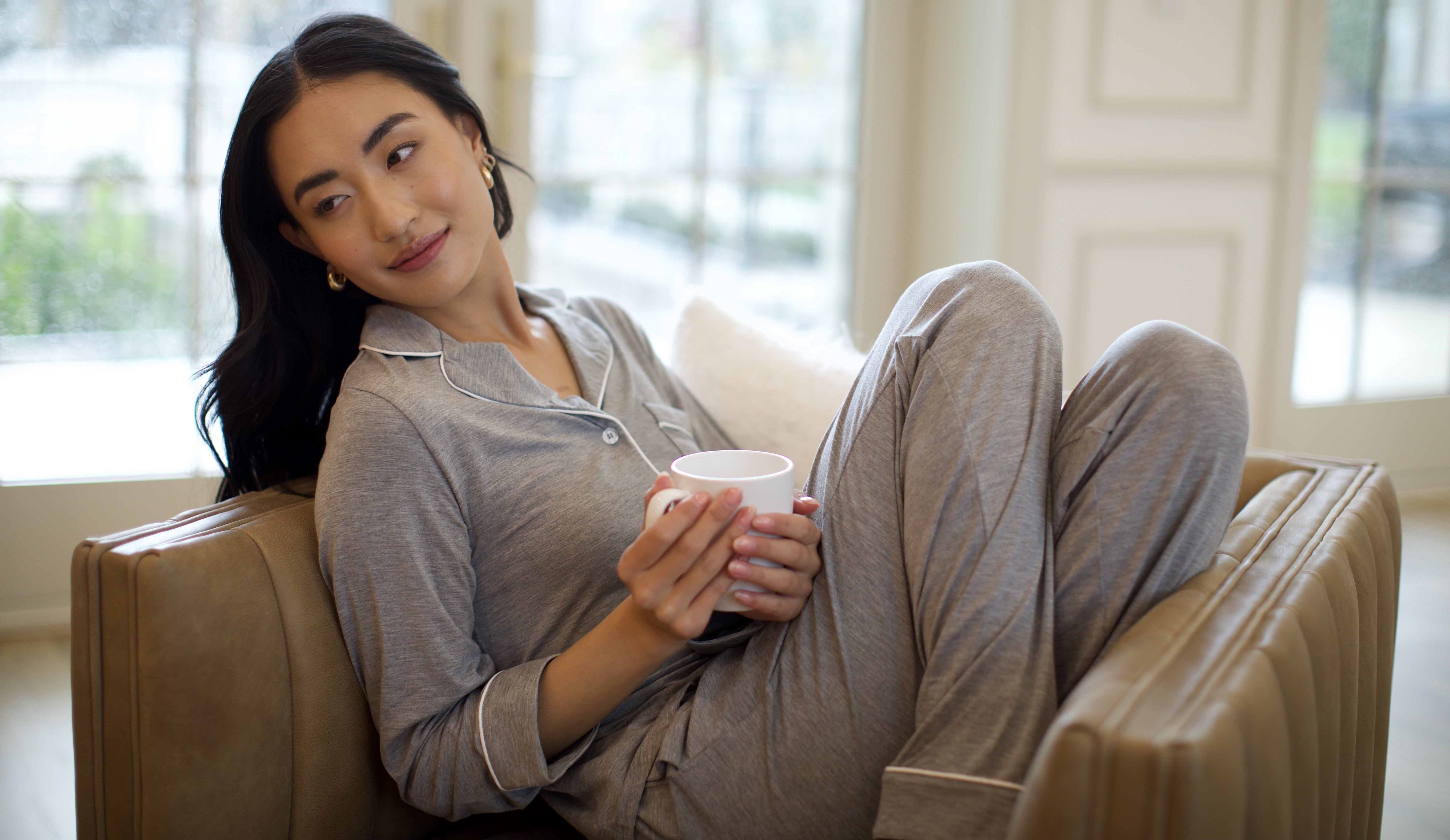 A woman in gray pajamas lounges on a couch, holding a mug and looking to the side with a relaxed, thoughtful expression. Soft natural light fills the cozy indoor setting.