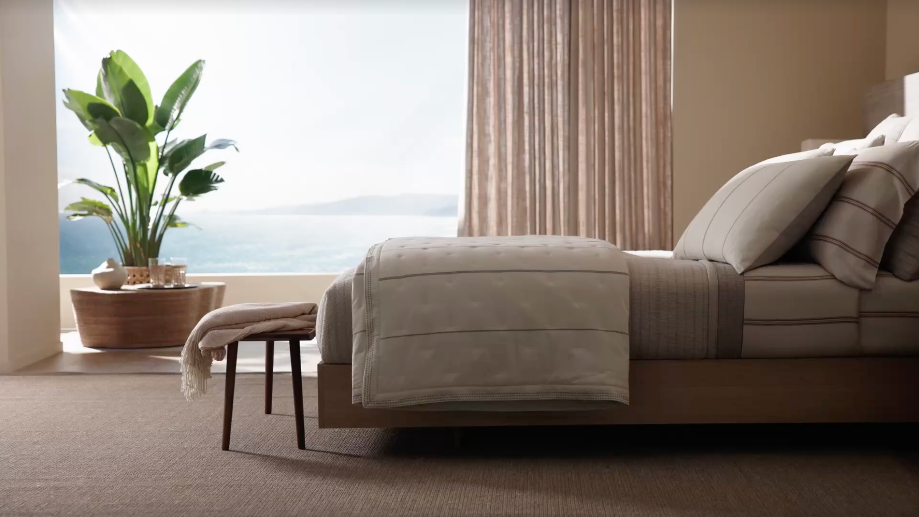 A neatly made bed with striped bedding sits by large windows overlooking the sea. A green potted plant and wooden bench with a blanket add warmth to the bright, minimalist bedroom.