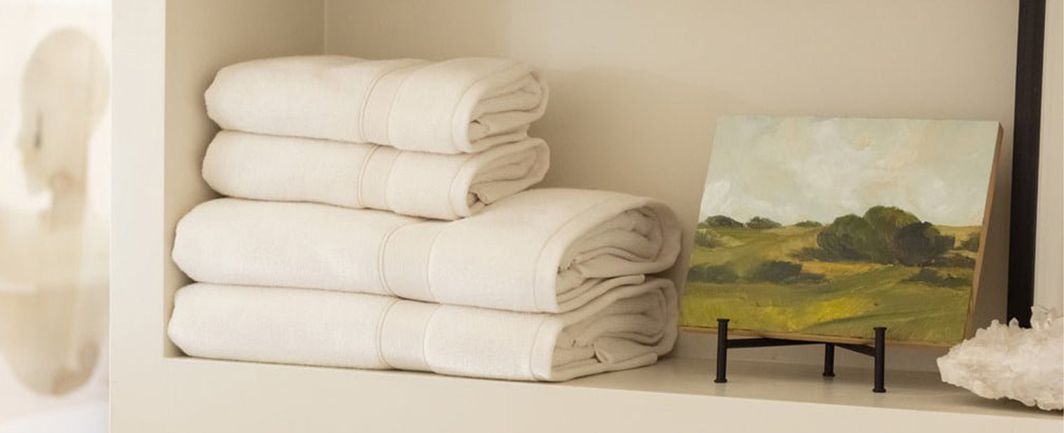 A stack of Cozy Earth Luxe Bath Towels sits on a shelf next to a landscape painting