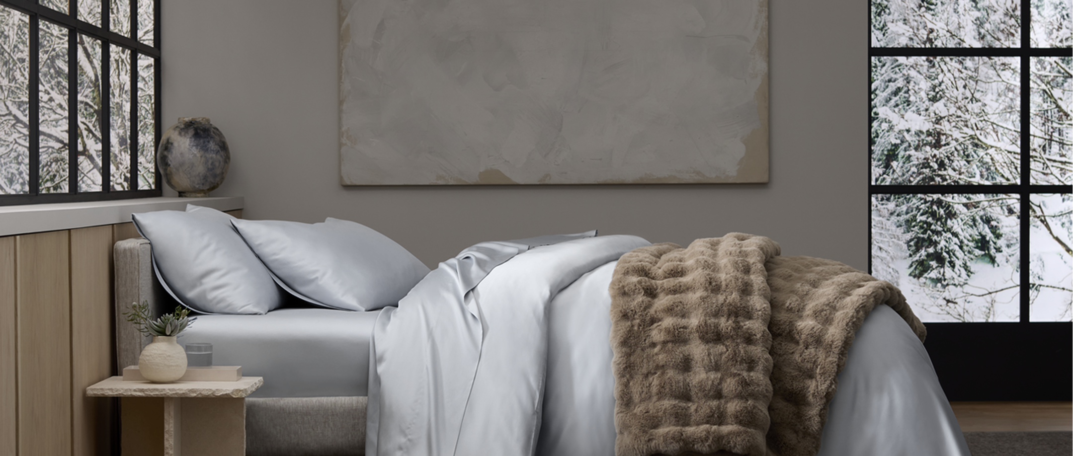 A neatly made bed with light gray sheets and pillows, a textured brown throw blanket, a small side table with decor, and large windows showing a snowy outdoor scene.