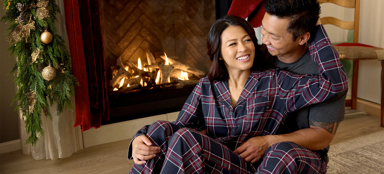 A couple in matching plaid pajamas sits smiling and embracing each other in front of a lit fireplace, with holiday decorations hanging nearby.