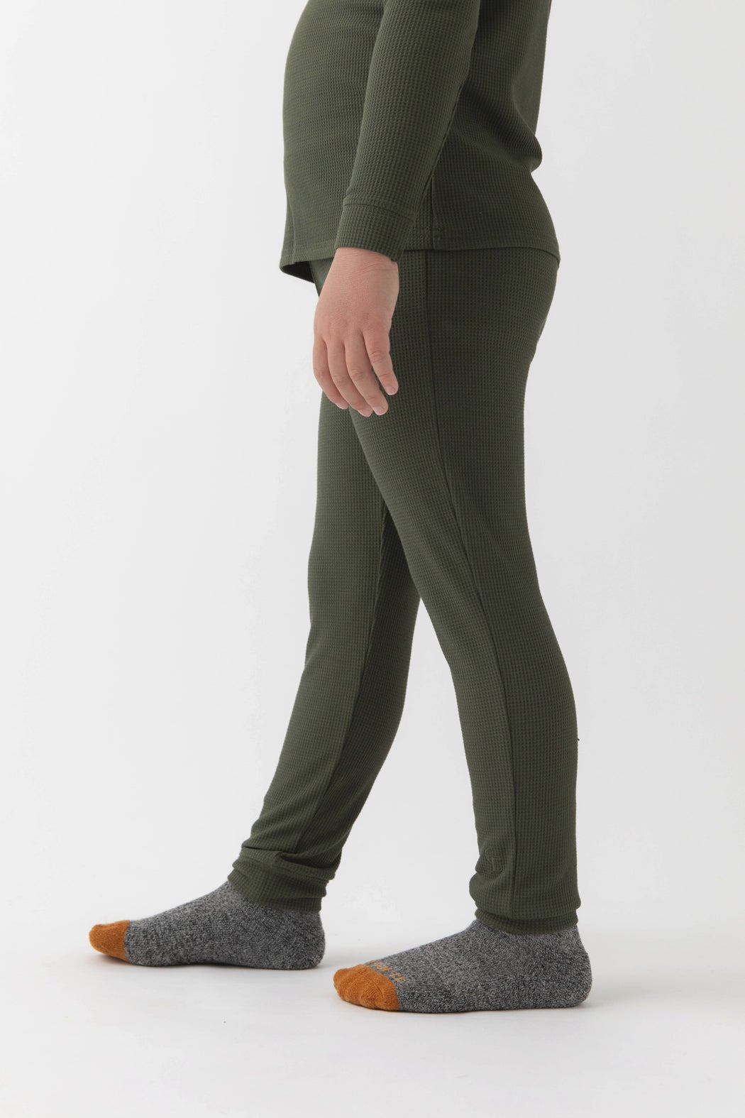 A child stands sideways on a white background, wearing Cozy Earth's Children's Bamboo Waffle Knit Base Layer Pant in green and a matching top. The child also has on gray socks with orange toes, and the face is not visible.