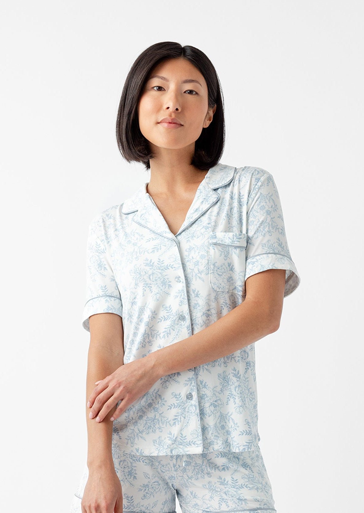 A person with shoulder-length black hair wears the Women's Bamboo Stretch-Knit Short Sleeve & Pant Pajama Set by Cozy Earth, featuring a light blue color and subtle floral pattern. They stand against a white background, looking slightly to the side. |Color:Sky Toile