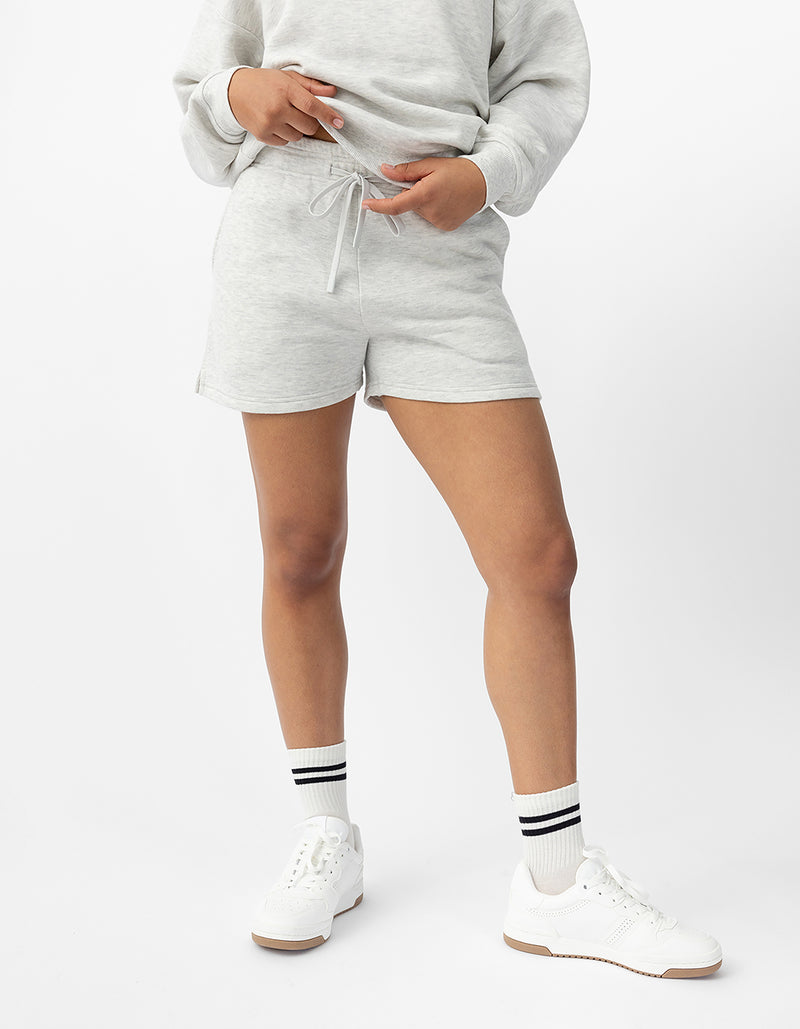 A person is wearing Cozy Earth's Women's CityScape Shorts paired with a light gray sweatshirt, white sneakers, and socks featuring black stripes. They stand against a plain white background with one hand pointing downwards.