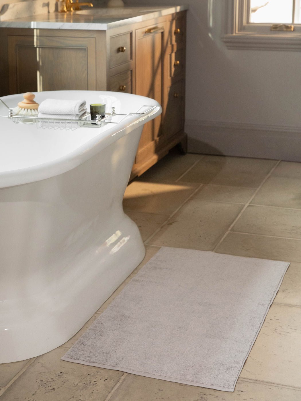 The Cozy Earth Loop Terry Bath Mat is shown on a tiled bathroom floor beside a freestanding tub, with folded towels and toiletries on a metal bath caddy. A wooden vanity with drawers appears in the background.