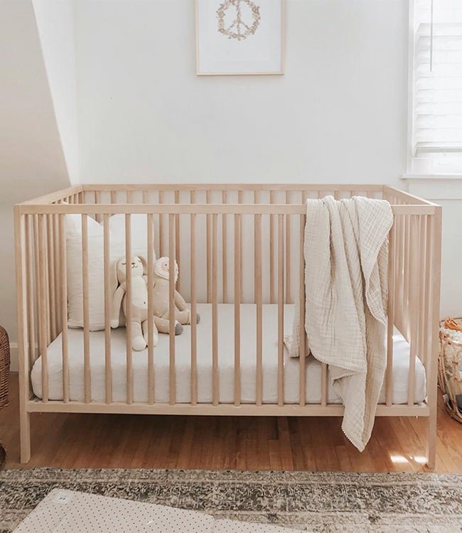 A minimalist nursery with a light wooden crib dressed in Cozy Earth’s Bamboo Fitted Sheet, white bedding, two stuffed animals, and a cream blanket draped over the side. The white-walled room features a window and framed print above the crib.
