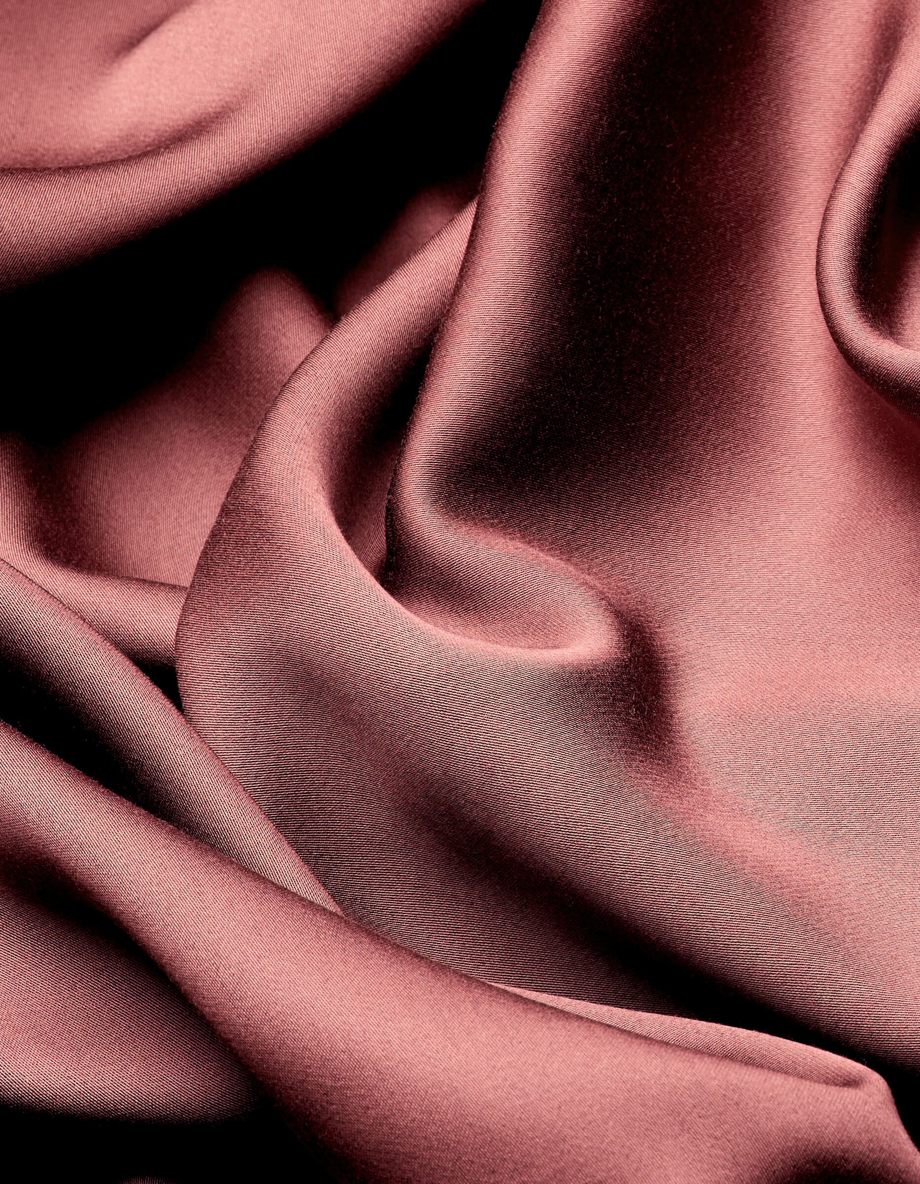 Smooth, shiny Bamboo Sheet Set by Cozy Earth drapes in soft, flowing folds, creating elegant highlights and shadows across the rose-colored satin surface.