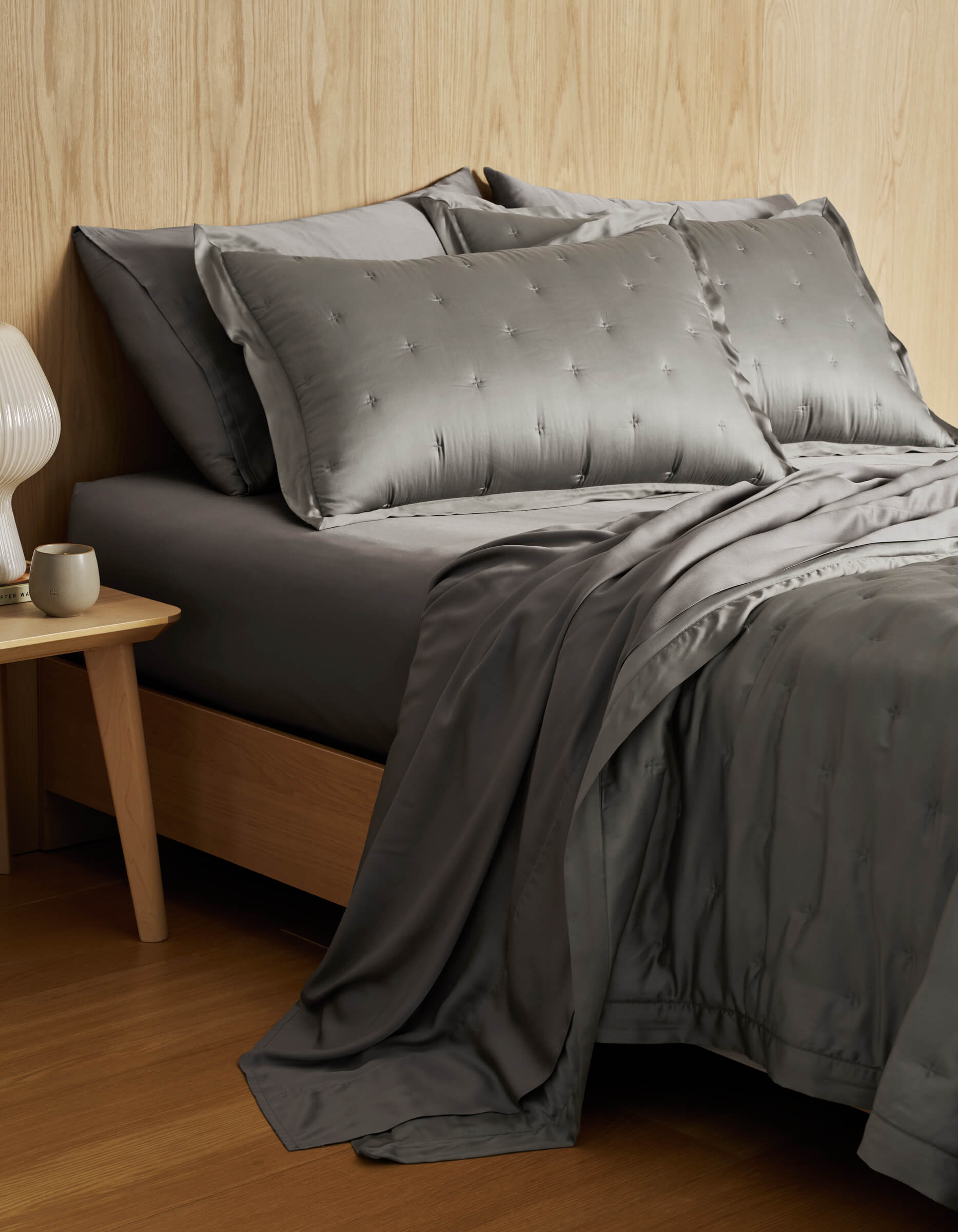A modern bedroom features a Cozy Earth Bamboo Quilt with gray silk bedding and matching pillows on a wooden bed frame, beside a wooden nightstand with a white ceramic lamp and cup, set against a light wood-paneled wall.|Color:Charcoal