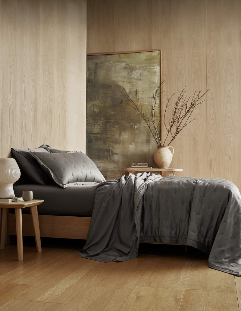 A modern bedroom with light wood walls and floors showcases a neatly made bed featuring the Cozy Earth Bamboo Quilt Bundle, complemented by a bedside table with a white lamp, a ceramic vase with bare branches, and a large abstract painting.