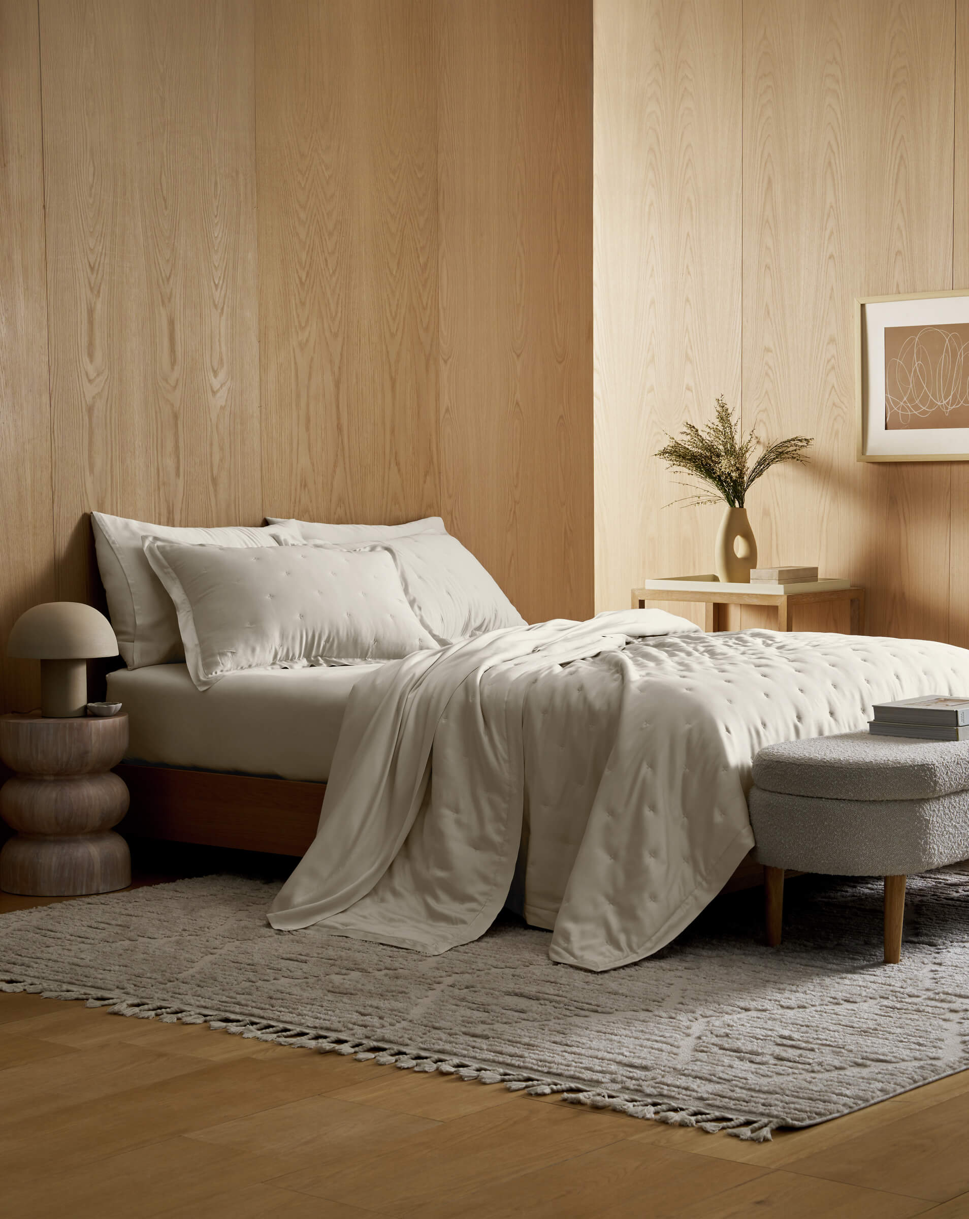 A minimalist bedroom features light wood-paneled walls, a bed dressed in Cozy Earth's Bamboo Quilt, white linens, a textured gray rug, modern bedside table with lamp and greenery in a vase, plus abstract art on the wall.