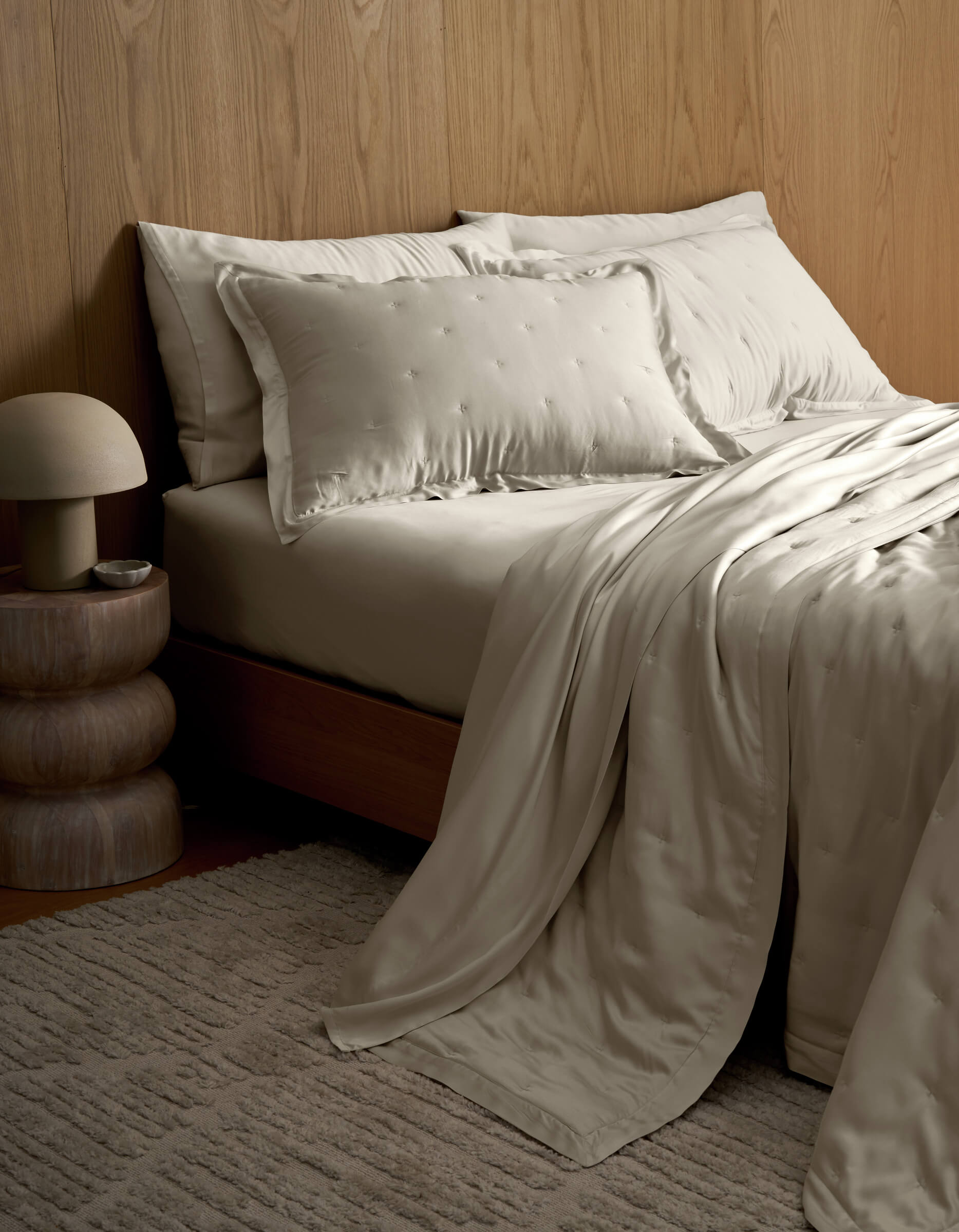 A modern bedroom with a neatly made bed dressed in the Cozy Earth Bamboo Quilt Bundle, set alongside a textured beige rug, a tiered wooden nightstand, and a small lamp against a wood-paneled wall.