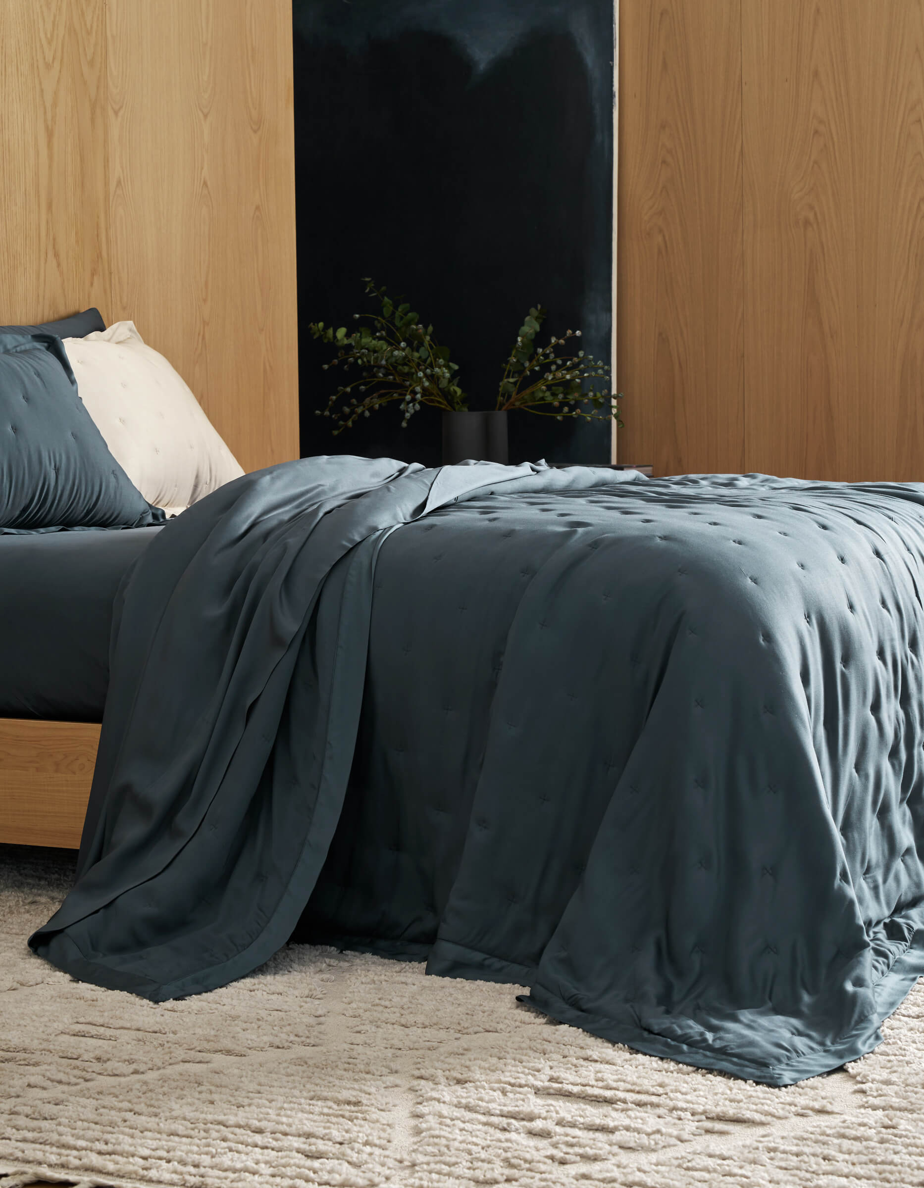 A neatly made bed features Cozy Earth's Bamboo Quilt in deep blue, paired with a textured blanket. Against a wooden wall, the room includes a light rug and a bedside table with a black vase of green foliage.