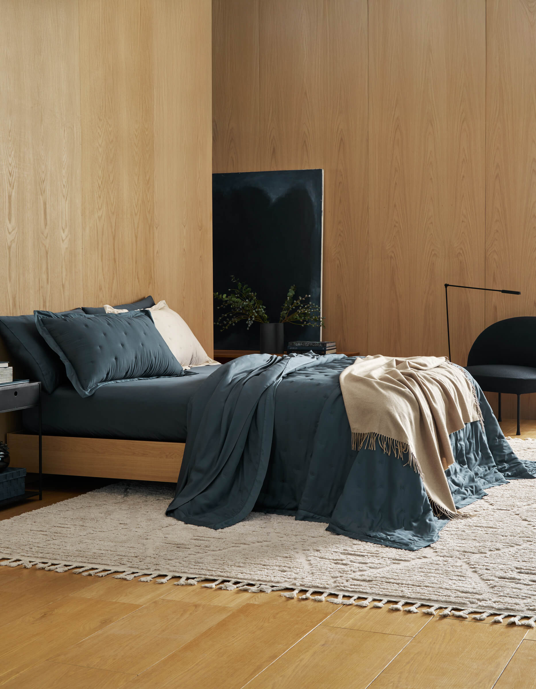 A modern bedroom features wood-paneled walls, a low bed with Cozy Earth's Bamboo Quilt Bundle in dark teal, a cream throw, and pillows. A neutral woven rug, black chair, side table, and minimalist decor create a cozy, stylish space.