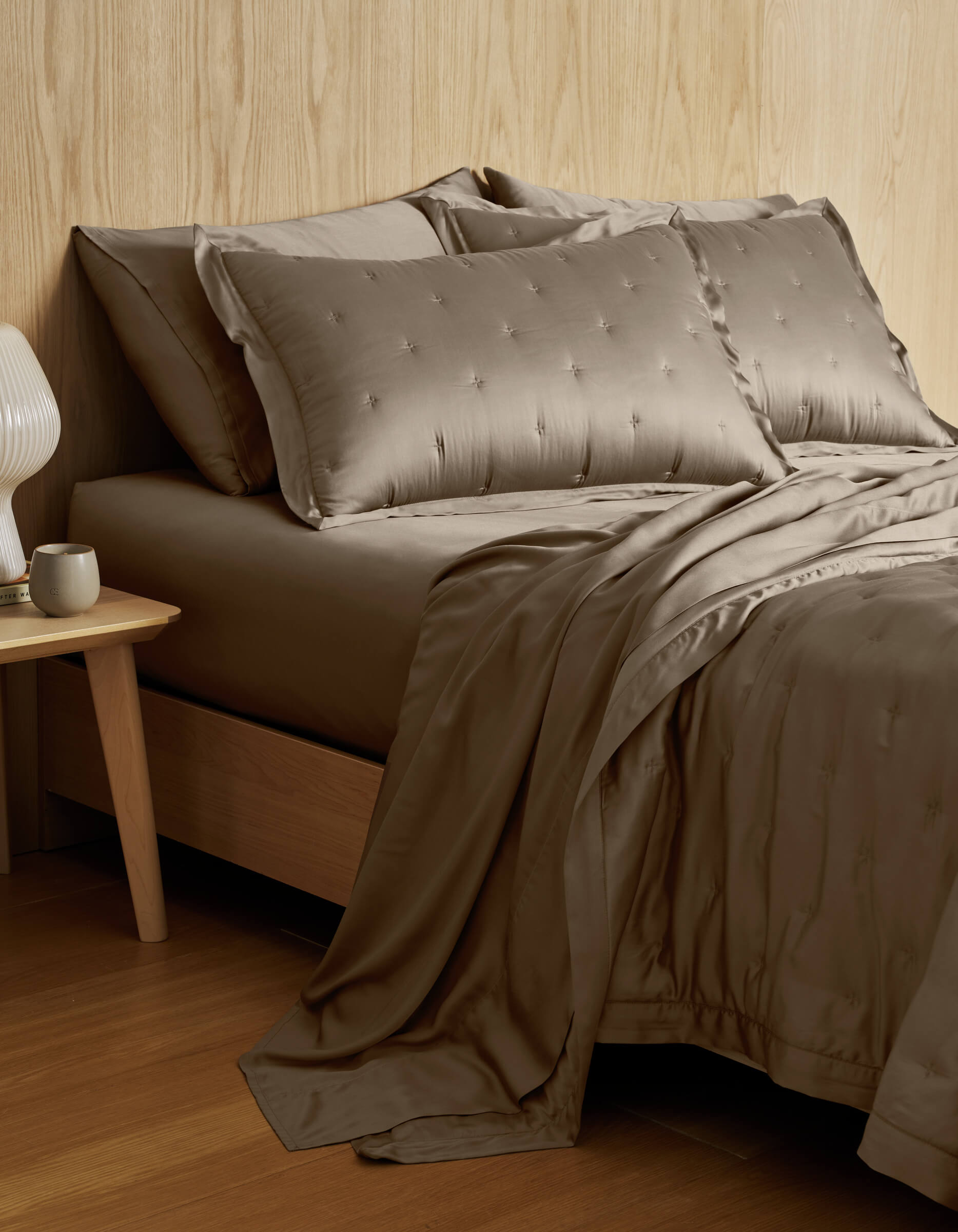 A neatly made bed with Cozy Earth’s Bamboo Quilt Bundle—taupe, embroidered bedding with matching pillows—beside a wooden nightstand holding a white lamp and coffee mug, set against a light wood-paneled wall.