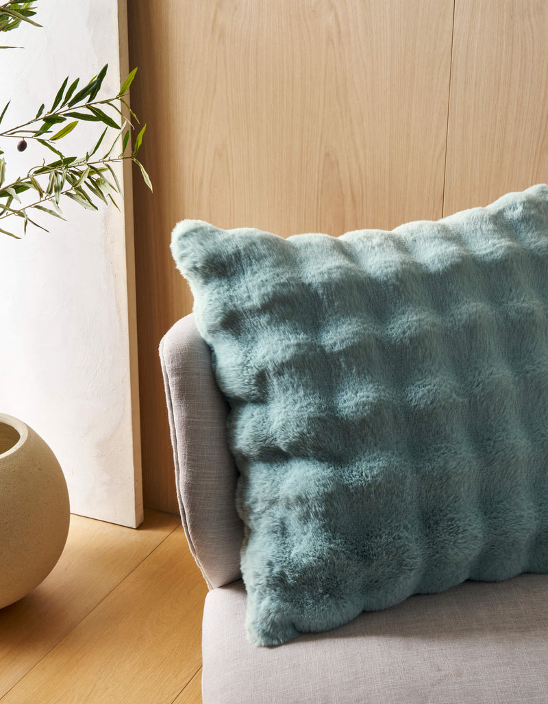 The Bubble Cuddle Pillow by Cozy Earth, featuring a soft textured light blue cover, sits on a light gray upholstered chair beside a round beige planter with greenery, all against a light wood-paneled wall.