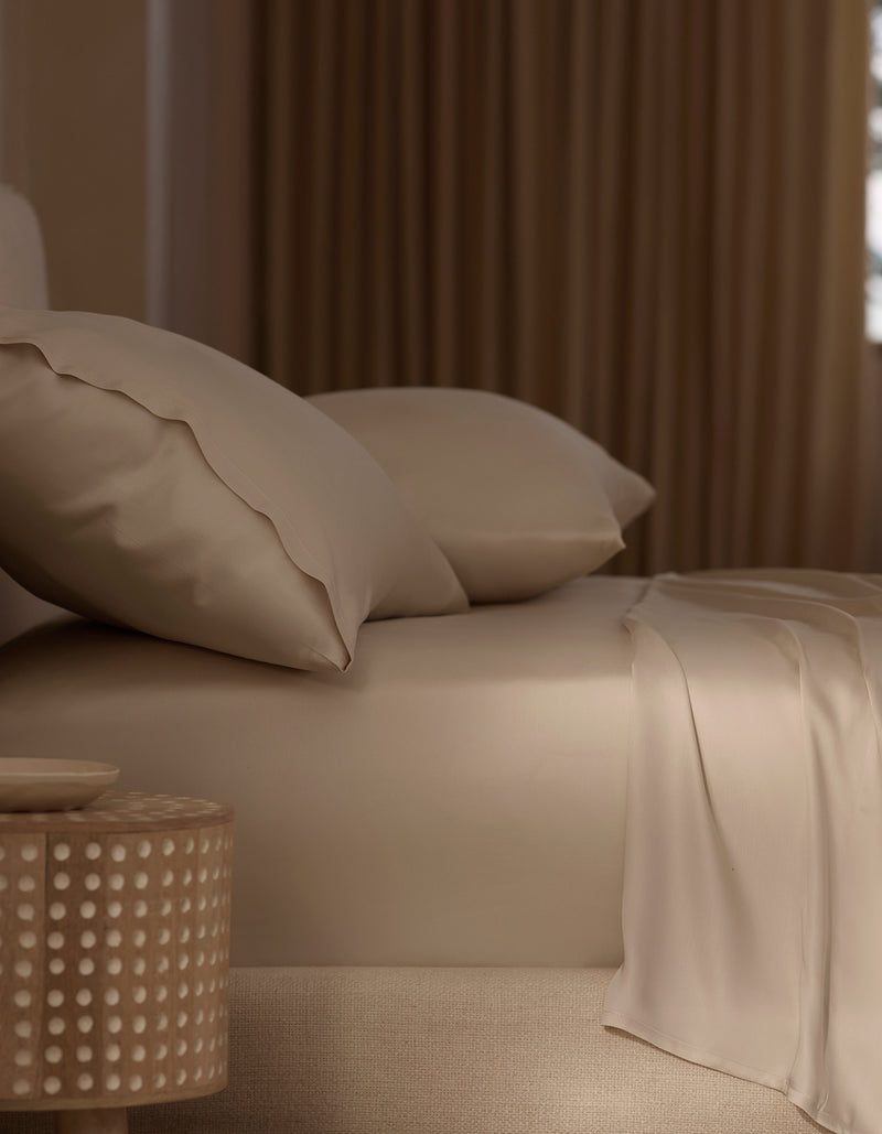A neatly made bed features Cozy Earth’s Bamboo Sheet Set in beige with matching pillows, beside a round wooden nightstand with a dotted design. Soft, warm light through curtains enhances the cozy atmosphere.
