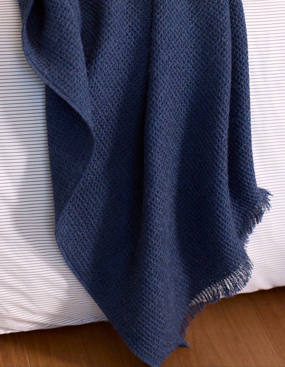 The Alpaca Waffle Throw by Cozy Earth, a textured dark blue blanket with fringed edges, drapes over a bed with white and gray striped sheets on a wooden floor.