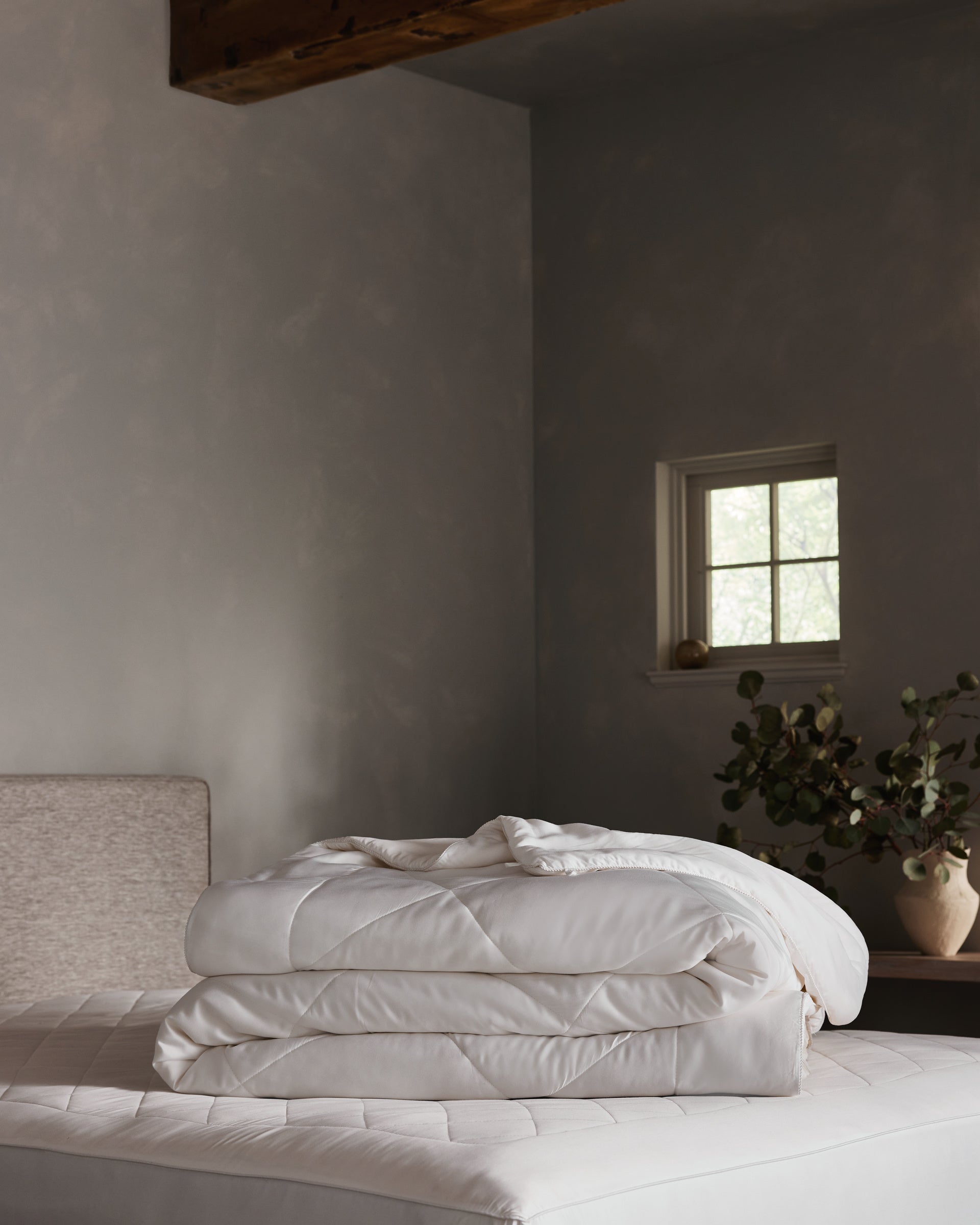 A neatly folded Cozy Earth Bamboo Viscose Comforter rests on a mattress in a softly lit bedroom with gray walls, a small window, and a vase with greenery atop a wooden nightstand.  Charcoal, Driftwood, Oat, Creme, White, Dove Grey, Light Grey, Shore, Pacific Blue, Peony, Walnut, Classic Stripe in Pacific, Classic Stripe in Ebony, Classic Stripe in Laurel, Mulberry, River