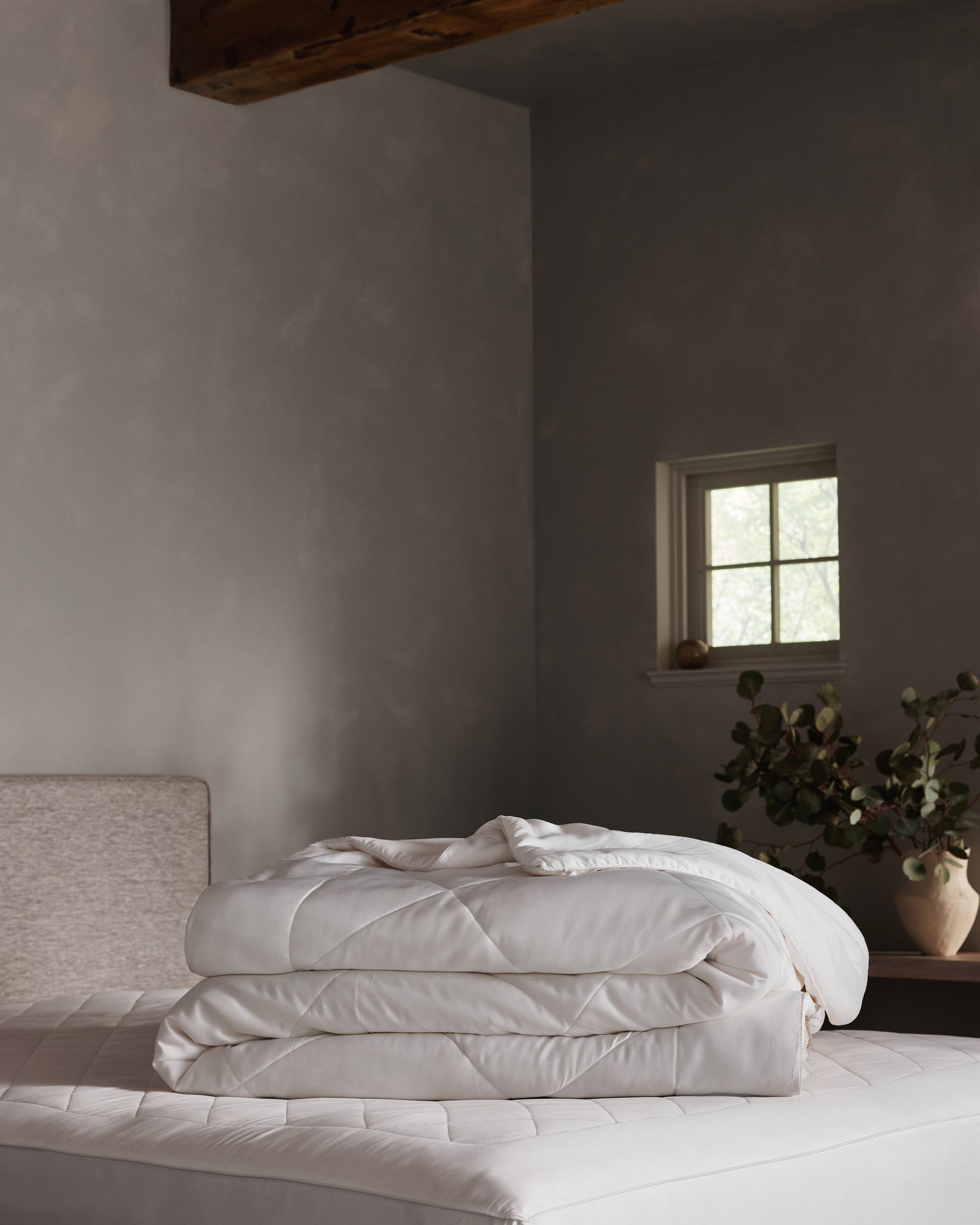 A neatly folded Cozy Earth Bamboo Viscose Comforter rests on a mattress in a softly lit bedroom with gray walls, a small window, and a vase with greenery atop a wooden nightstand. 
