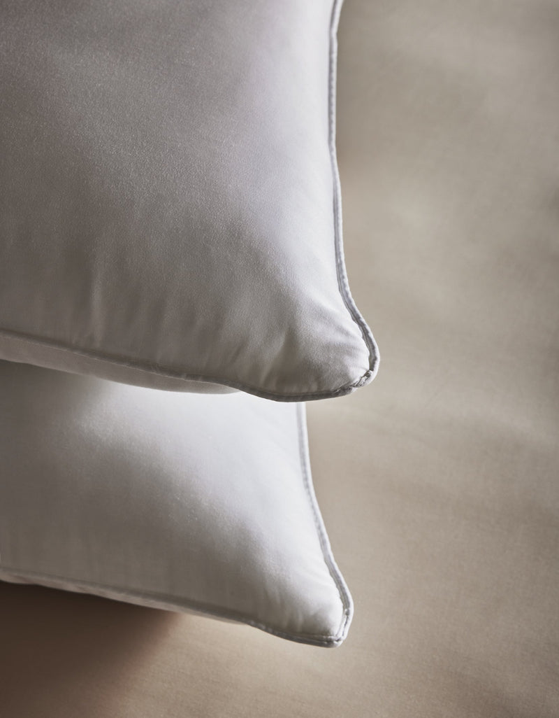 Two Bamboo Down Alternative Pillows by Cozy Earth with smooth, satiny pillowcases are neatly stacked on a light beige surface and viewed close up from the corner.Standard King