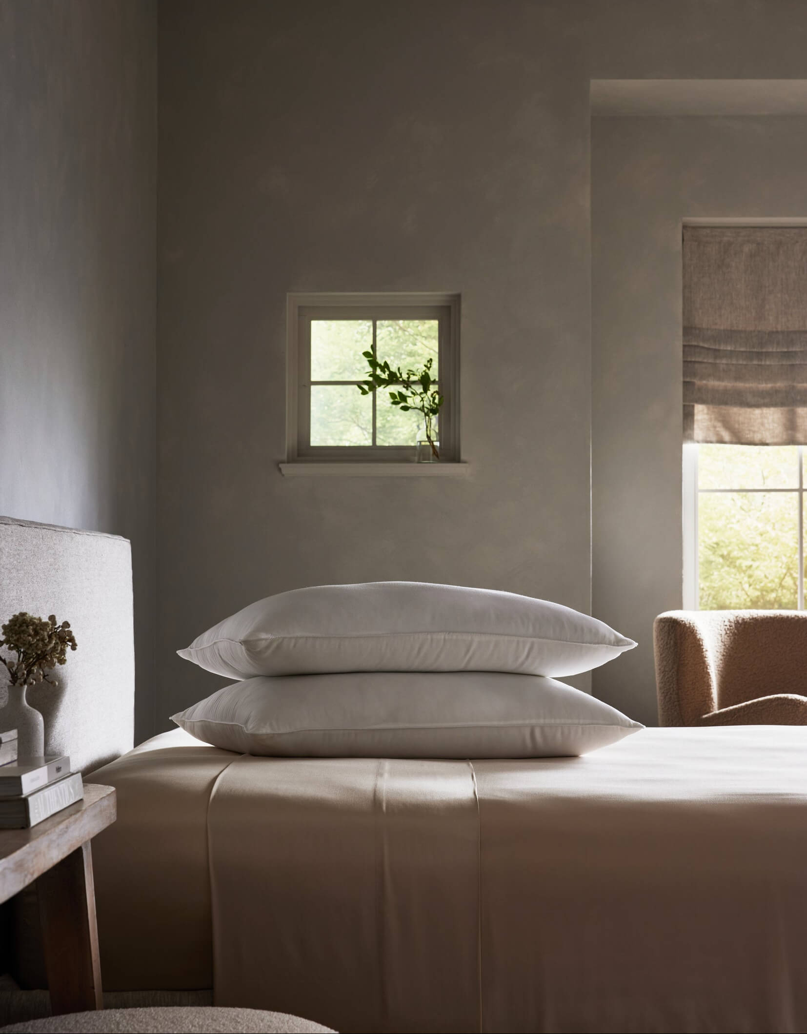 A minimalist, calming bedroom features Cozy Earth's Bamboo Insert Bundle—beige sheets with two stacked white pillows—by a small window with a vase of greenery, bathed in soft natural light.