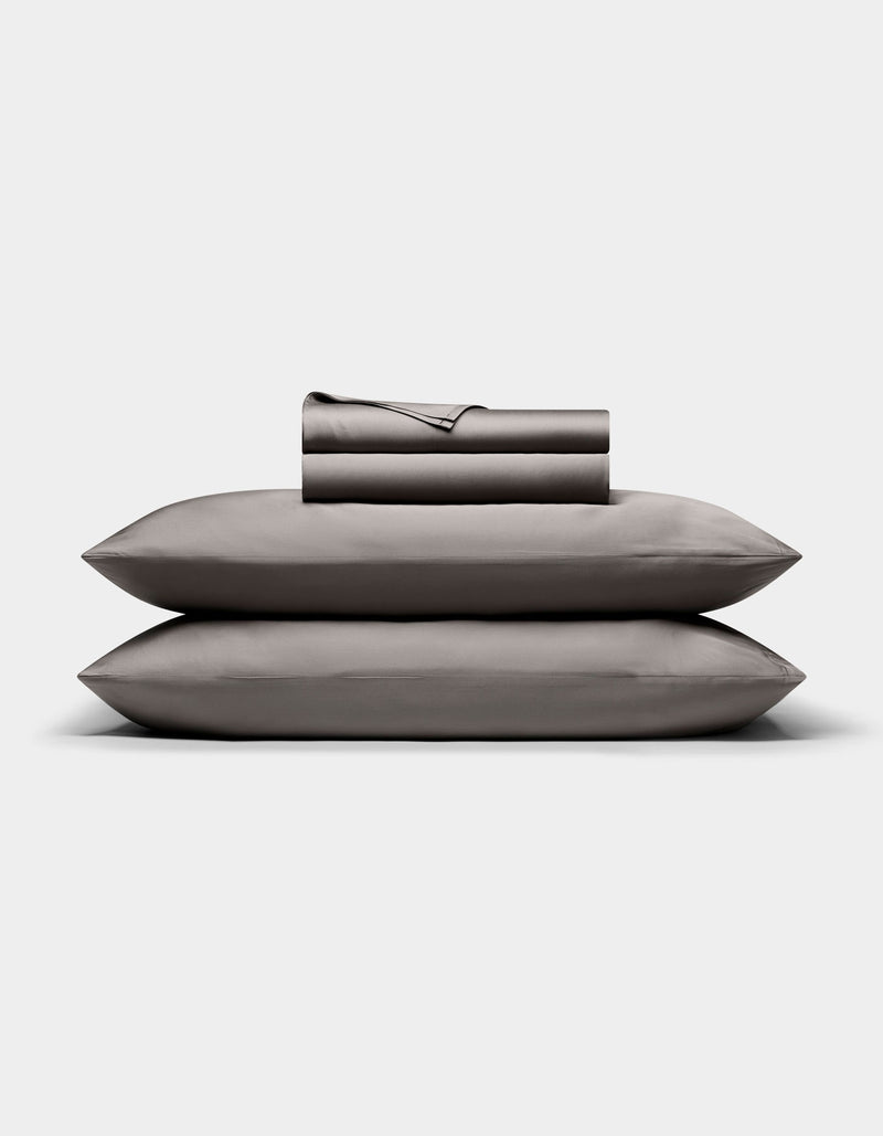 The Cozy Earth Bamboo Sheet Set, in dark gray, is neatly folded with matching pillowcases stacked atop two pillows, all arranged against a plain white background.