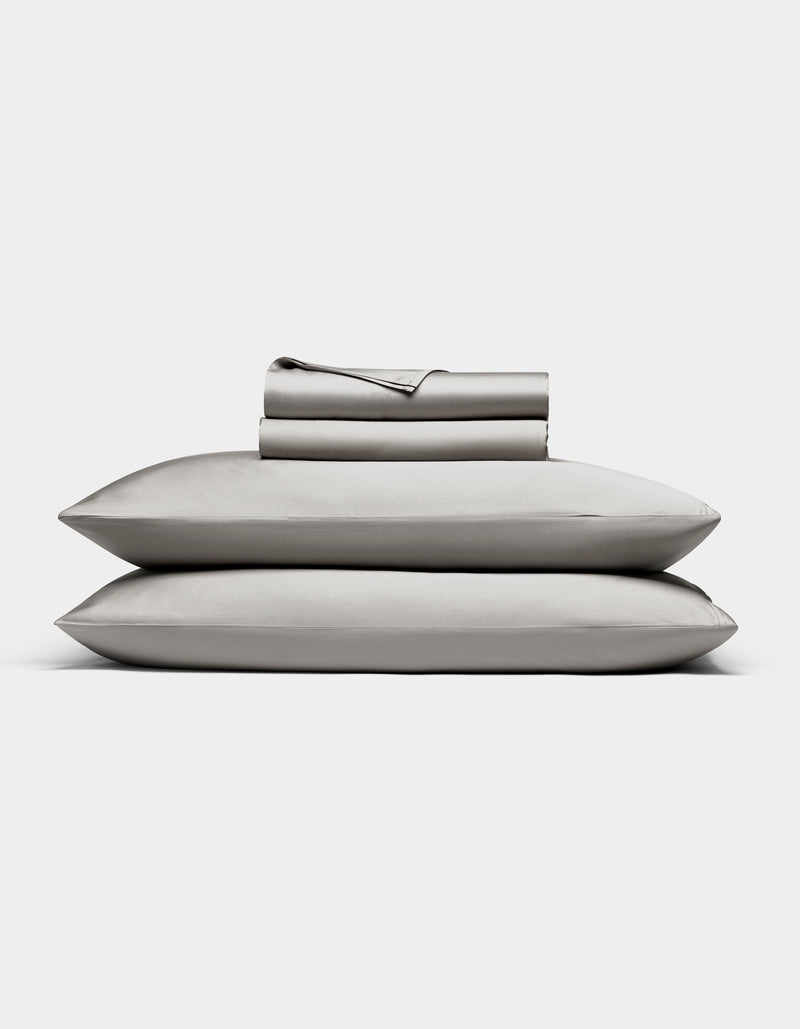The Bamboo Sheet Set by Cozy Earth in light gray is neatly folded, with two pillows stacked and both flat and fitted sheets rolled on top, all displayed against a white background.