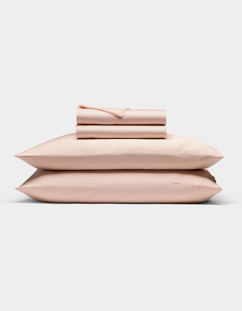 The Cozy Earth Bamboo Sheet Set in light pink is neatly folded with matching pillowcases, stacked on two pillows against a plain white background.