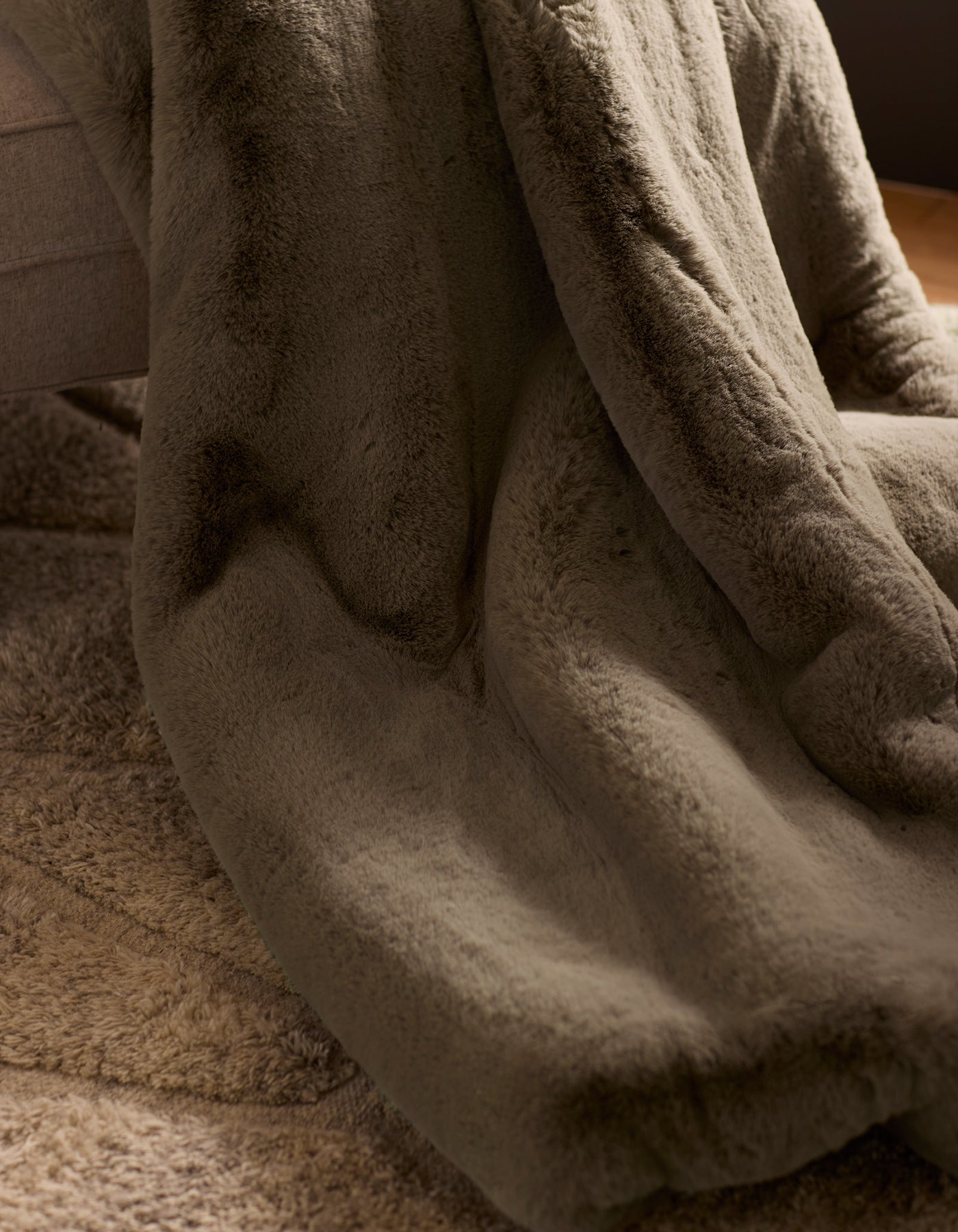 The Cozy Earth Cuddle Blanket, a soft plush brown faux fur throw, is draped over a couch edge and rests on a textured beige carpet in a warmly lit, cozy room.