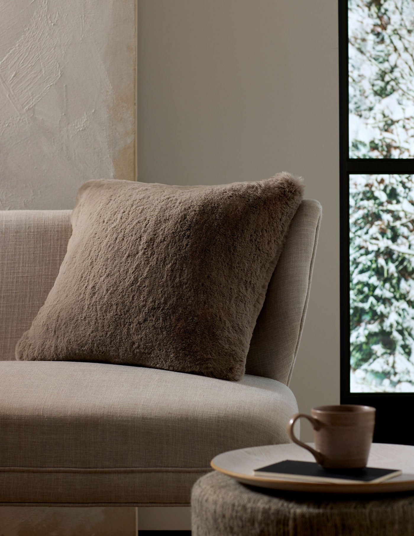 A cozy, neutral-toned living space features the Cozy Earth Cuddle Pillow in taupe on a beige sofa next to a round side table with a ceramic mug and book, framed by a window view of snowy trees.|Color:Walnut |Size:24" x 24"