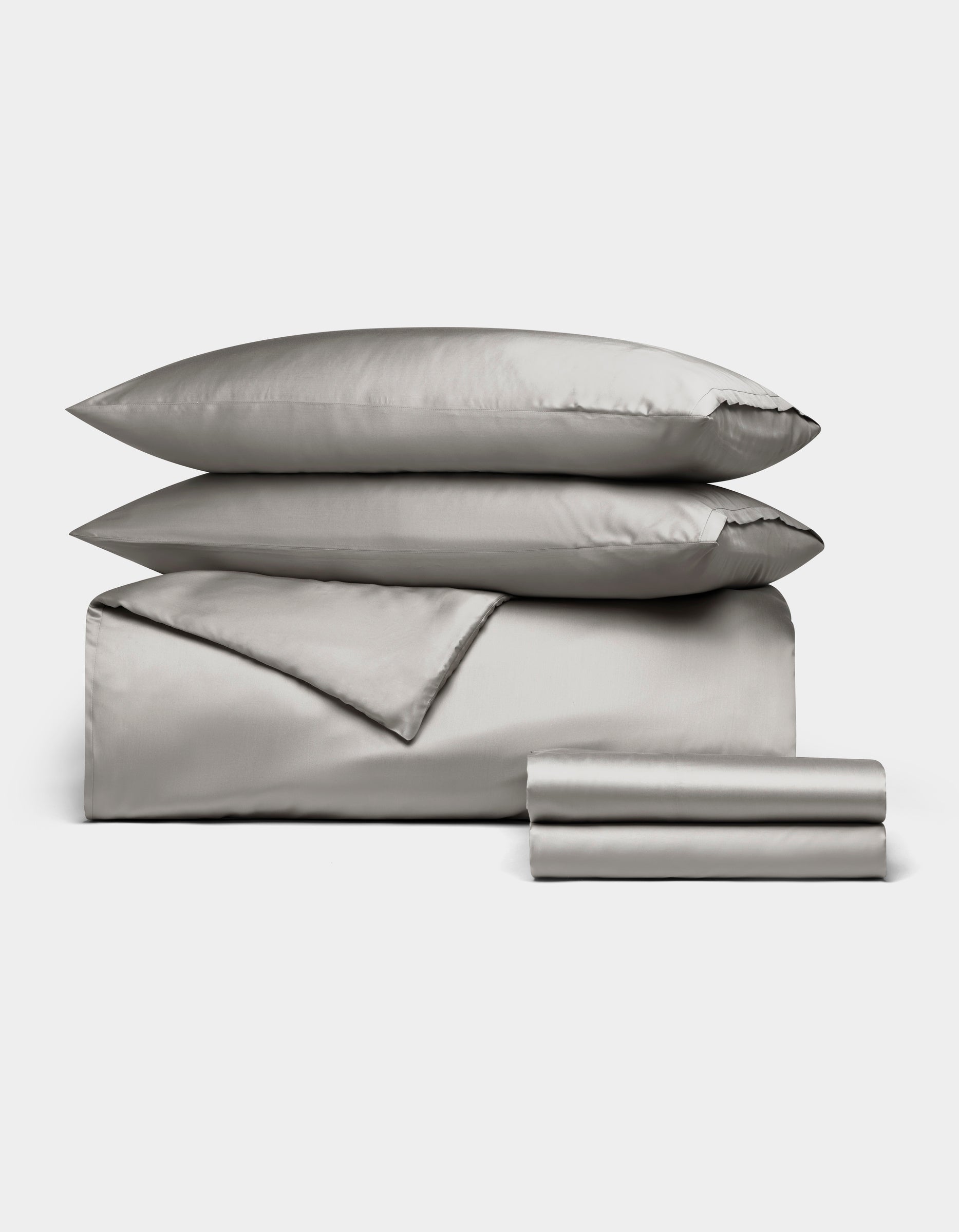 The Cozy Earth Bamboo Bedding Deluxe Bundle features light gray satin bedding with two pillows, a folded duvet, and stacked sheets, all neatly arranged on a white background.|Color:Light Grey