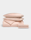 The Cozy Earth Bamboo Bedding Deluxe Bundle, featuring two pillows, a comforter, and two folded sheets in pale pink, is neatly stacked and displayed against a plain white background.|Color:Peony