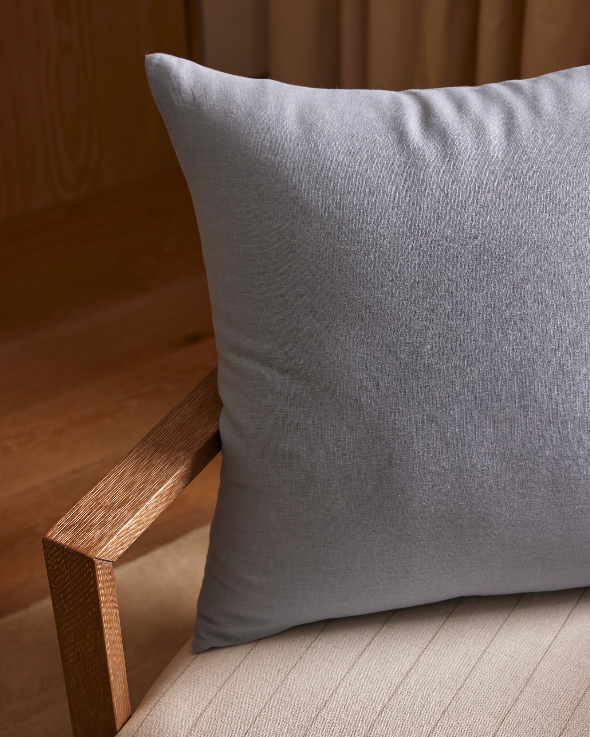 The Cozy Earth Linen Throw Pillow, in light gray, sits on a beige upholstered chair with a wooden armrest, set against a backdrop featuring wood accents and curtains. 