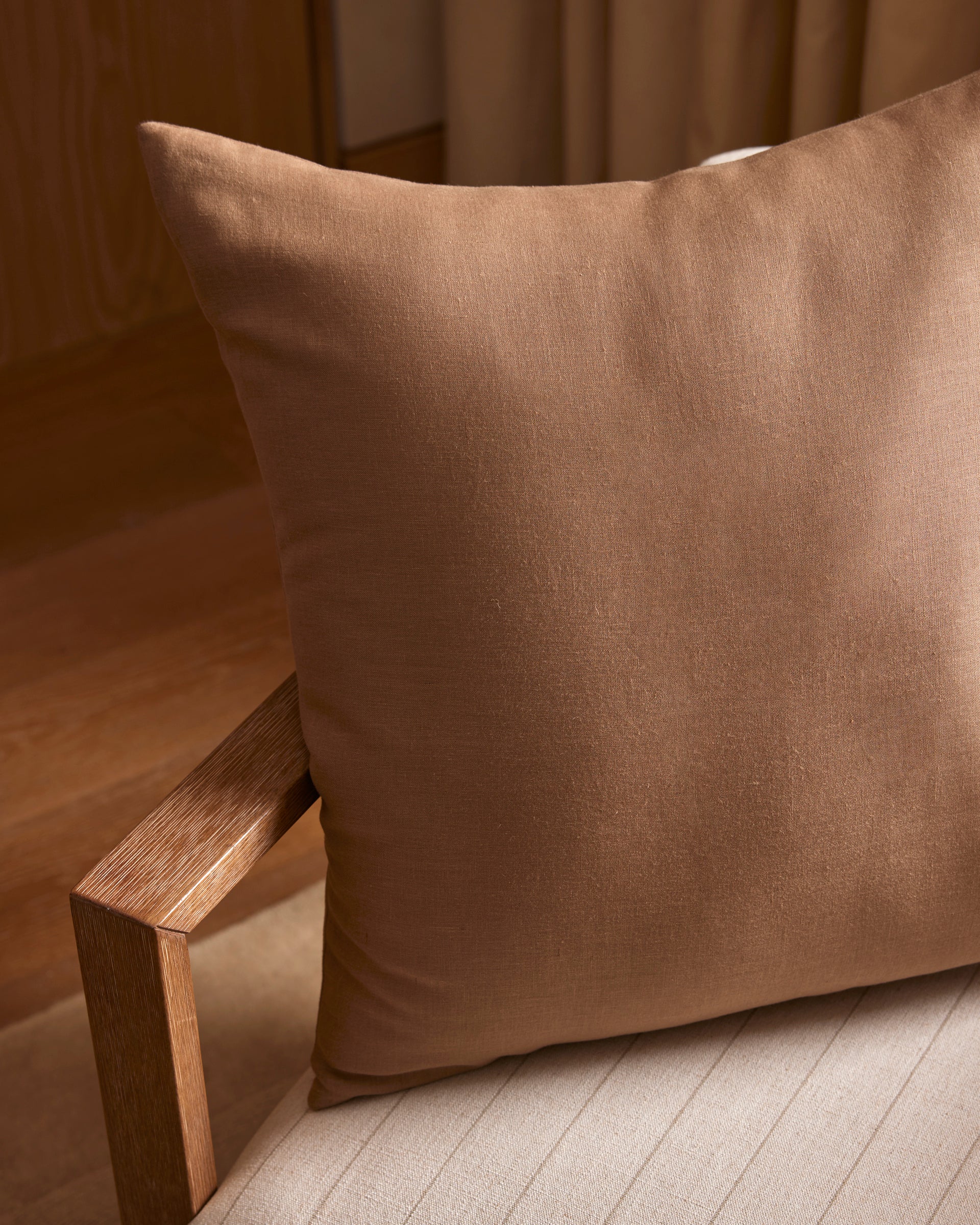 A Linen Throw Pillow by Cozy Earth sits on a beige, striped cushioned chair with a wooden armrest, in a warmly lit room featuring wooden floors and neutral-colored walls.