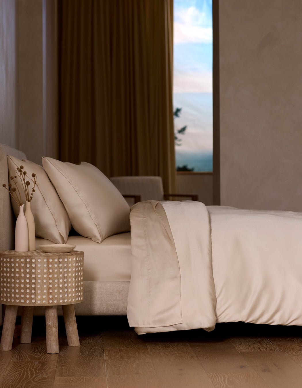 A neatly made bed with the Cozy Earth Bamboo Sheet Set in cream sits next to a round wooden nightstand holding two vases of dried flowers, as soft natural light streams through a window with beige curtains.