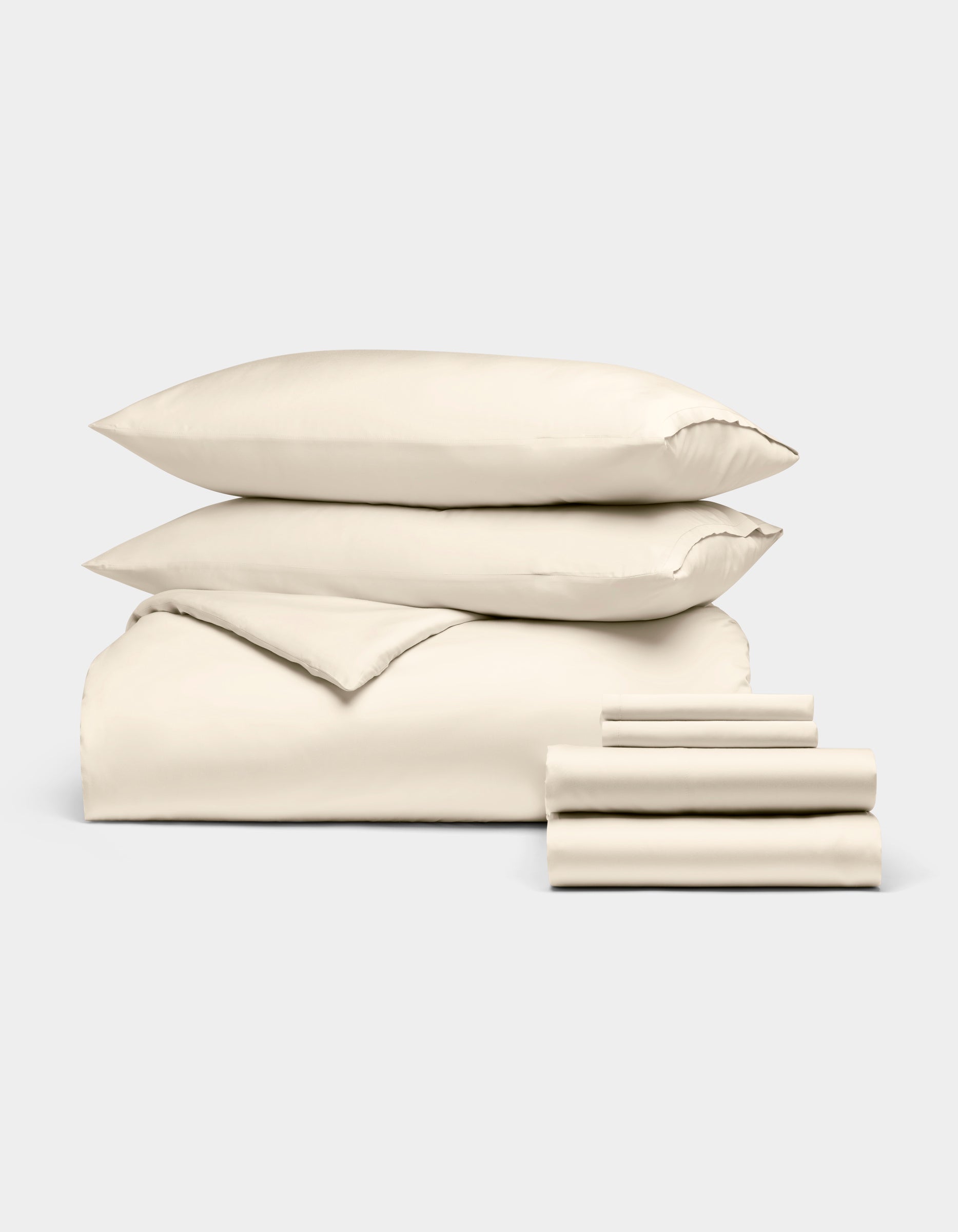 The Bamboo Bedding Ultimate Bundle by Cozy Earth, shown on a light grey background, features a neatly folded cream-colored duvet, two pillows, and a stack of sheets.|Color:Creme