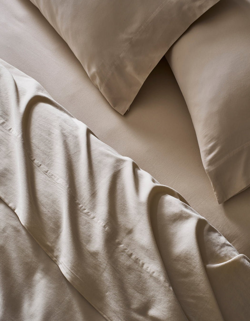 A close-up of a neatly made bed features Cozy Earth's Soft-Wash Cotton Sheet Set in beige. The slightly rumpled sheets and pillowcases enhance the soft, textured look of the smooth fabric.