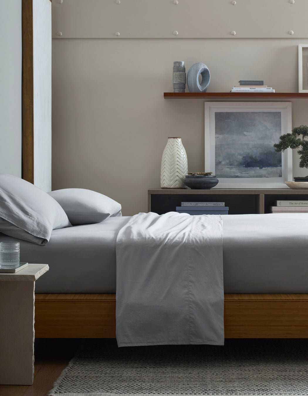 A neatly made bed with Cozy Earth Soft-Wash Cotton Pillowcases sits beside a nightstand with a glass of water. Behind the bed, shelves display books, vases, framed art, and a bonsai, creating a calm, modern bedroom.