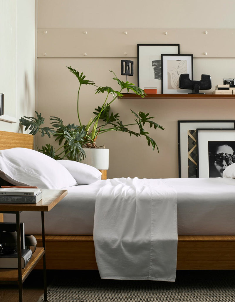 A modern bedroom features Cozy Earth's Soft-Wash Cotton Flat Sheet, a wooden bed with white bedding, a leafy plant in a white pot, framed art and decor on a shelf above the bed, and a side table with books.