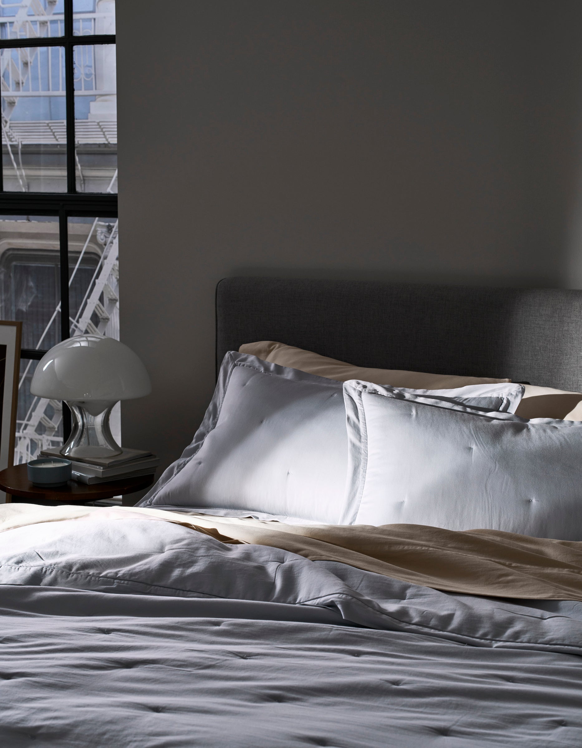 A bed with Cozy Earth Soft-Wash Cotton Quilted Shams in white and beige sits by a sunlit window. A bedside table holds a white lamp and books, while part of a gray upholstered headboard is seen.
