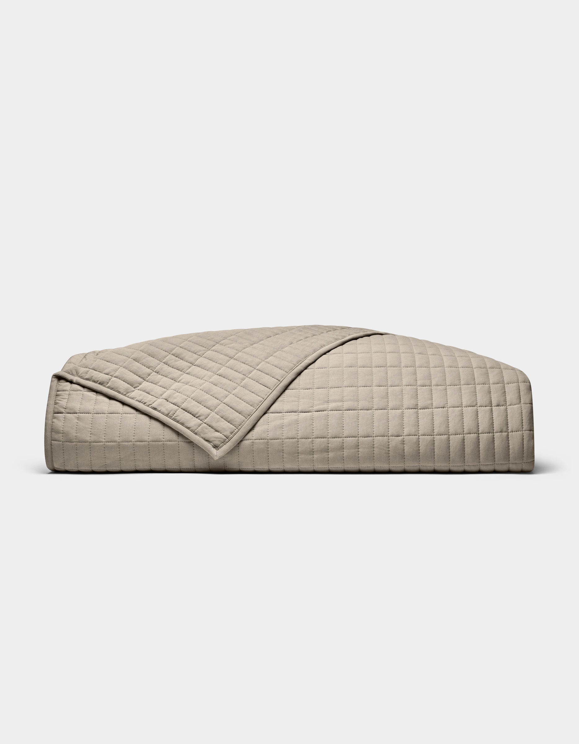 The Cozy Earth Bamboo Jersey Quilt, featuring a beige, quilted grid pattern, is neatly folded on a white background. A turned-down corner highlights its soft, luxurious texture.