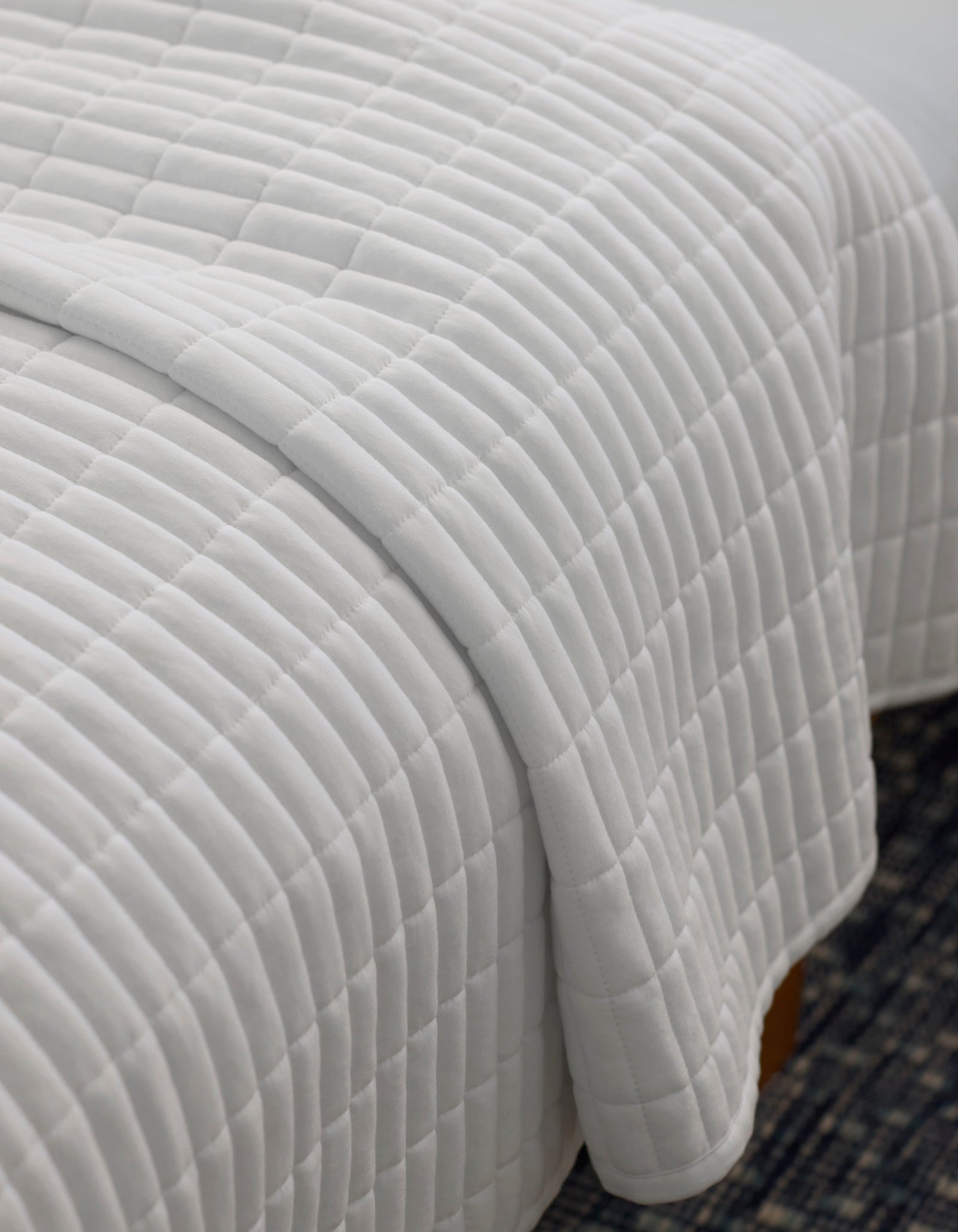 A close-up of a neatly made bed with the Cozy Earth Bamboo Jersey Quilt, featuring a white, square-patterned design, draped over the edge atop a dark patterned rug.