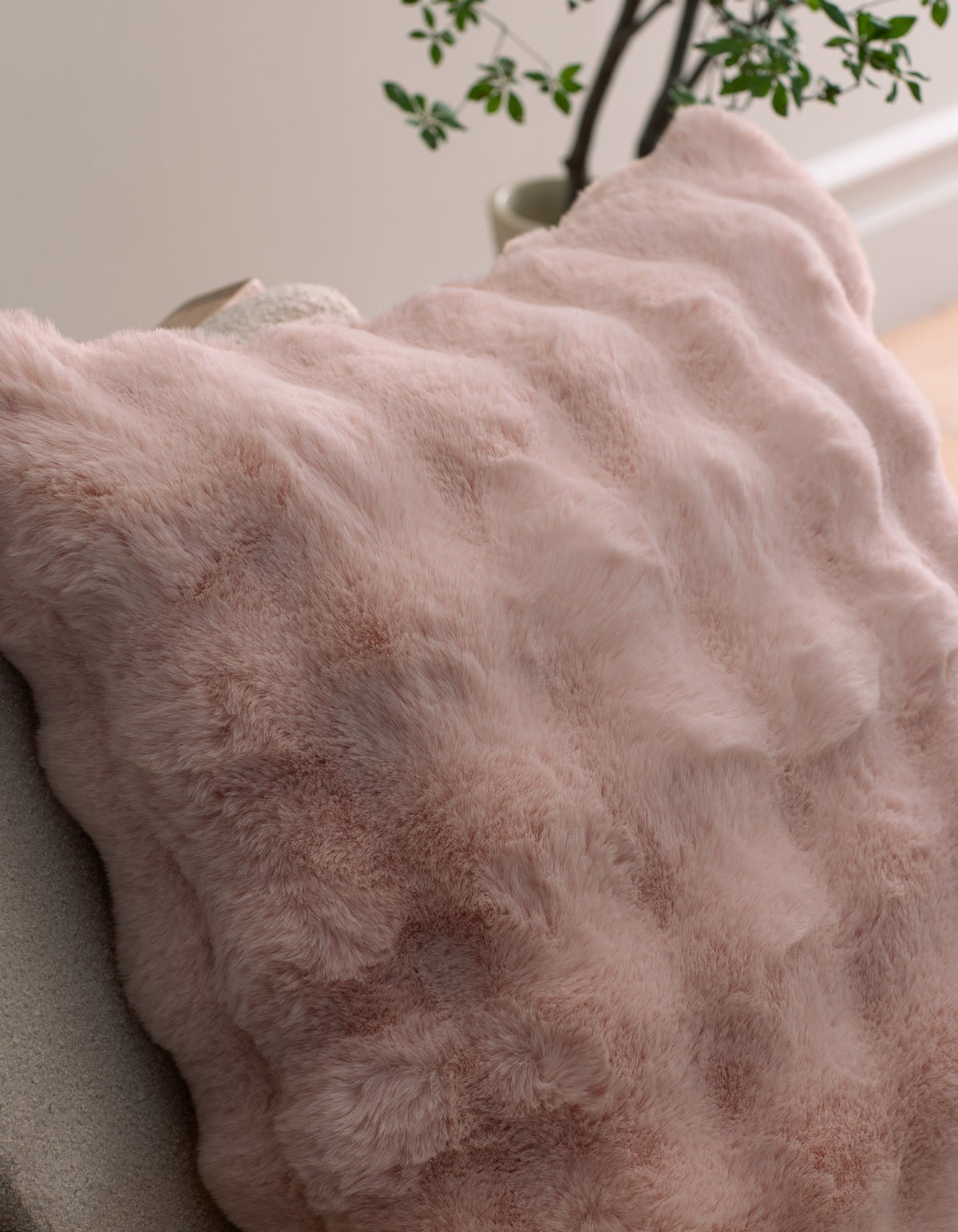 The Bubble Cuddle Pillow by Cozy Earth, featuring soft pink faux fur, sits on a light chair with a green potted plant visible in the background.