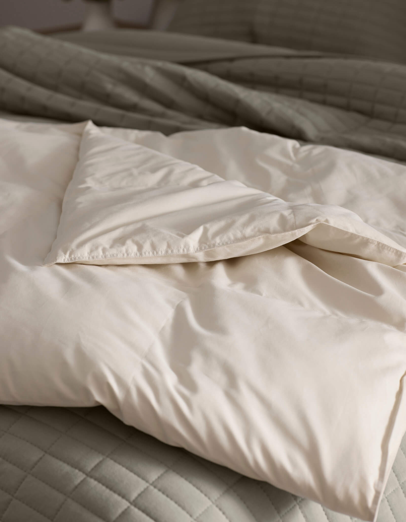 A close-up of Cozy Earth's Down Alternative Throw, folded atop a quilted light gray bedspread. The soft, cream-colored fabric looks cozy and inviting, with natural light highlighting its smooth texture.