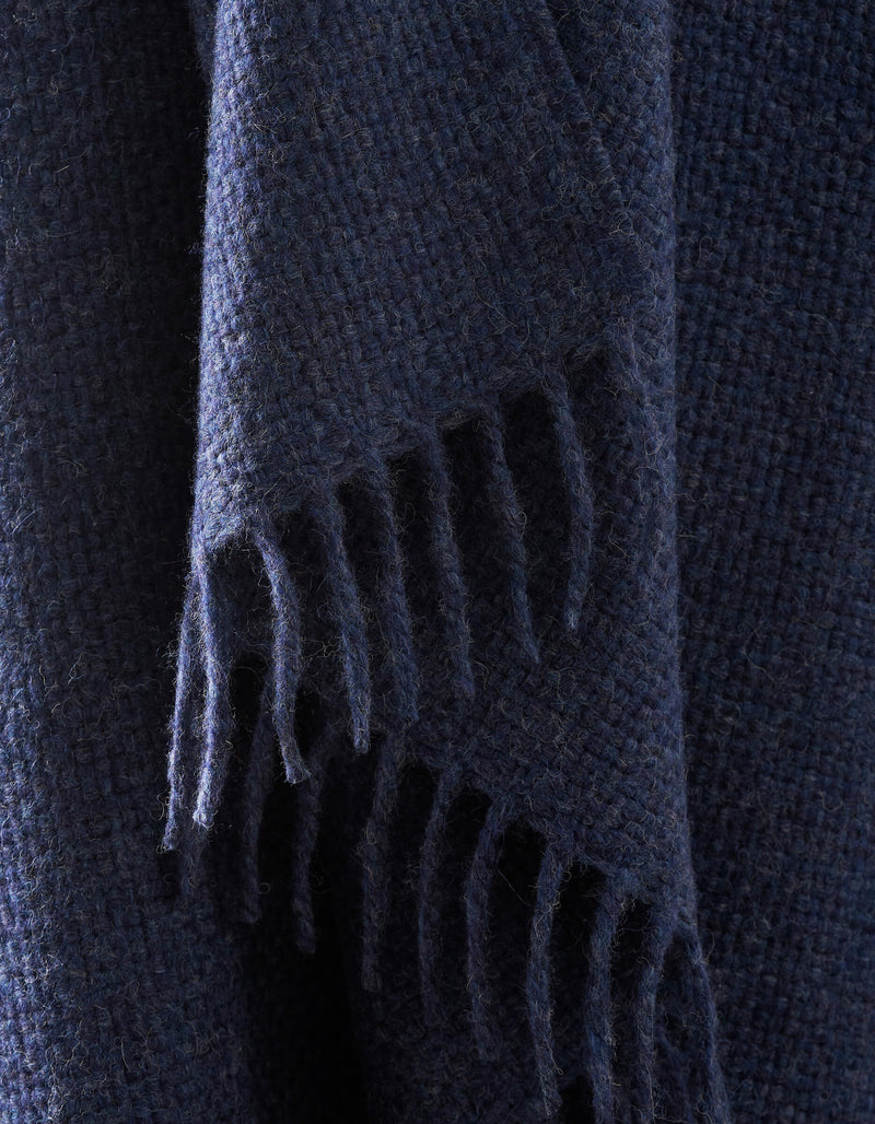 A close-up of the Lucia Throw by Cozy Earth shows its dark blue, textured woven fabric with fringed edges hanging down.