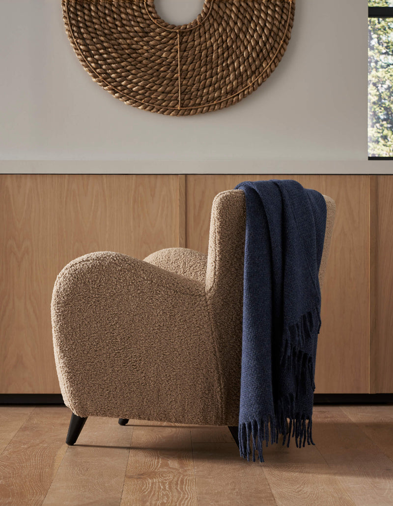 A cozy beige armchair with a Cozy Earth Lucia Throw draped over one arm sits on a wooden floor before light wood cabinets. Above the chair hangs a large, round, woven wall art piece.